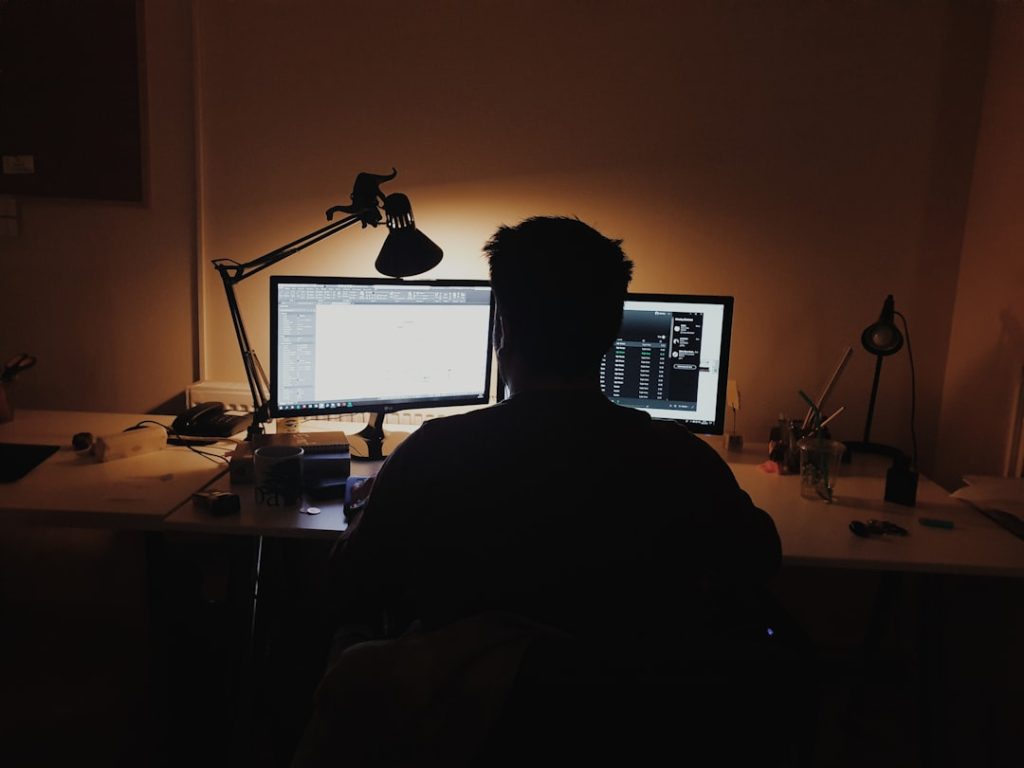 A lone founder working late at a dual-monitor desk in a dimly lit room, running outbound sales solo instead of hiring an SDR team.