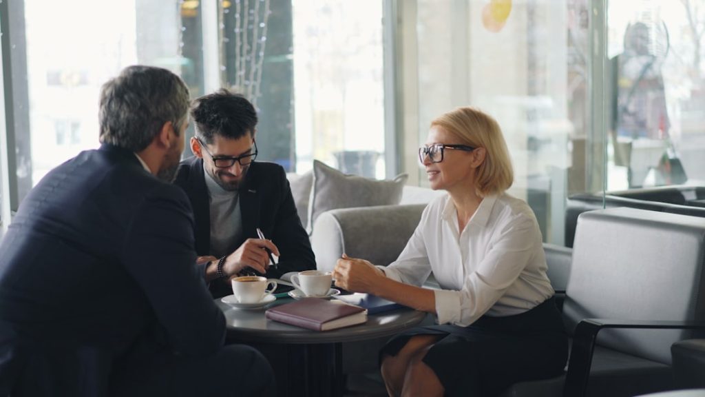 Three colleagues in business clothes talking around a small round table in a bright glass-walled lobby, with coffee cups, a closed notebook and a pen between them.
