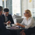 Three colleagues in business clothes talking around a small round table in a bright glass-walled lobby, with coffee cups, a closed notebook and a pen between them.