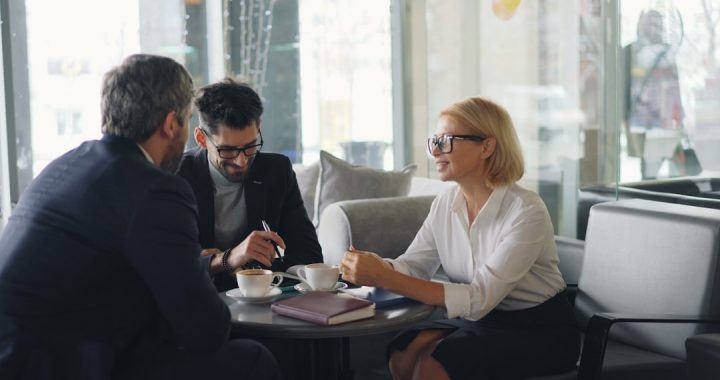 Three colleagues in business clothes talking around a small round table in a bright glass-walled lobby, with coffee cups, a closed notebook and a pen between them.