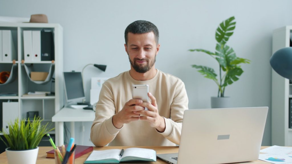 A solo founder sitting at a desk checking his phone, with a laptop and an open notebook beside him, scanning LinkedIn engagement signals before writing outreach messages.