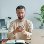 A solo founder sitting at a desk checking his phone, with a laptop and an open notebook beside him, scanning LinkedIn engagement signals before writing outreach messages.