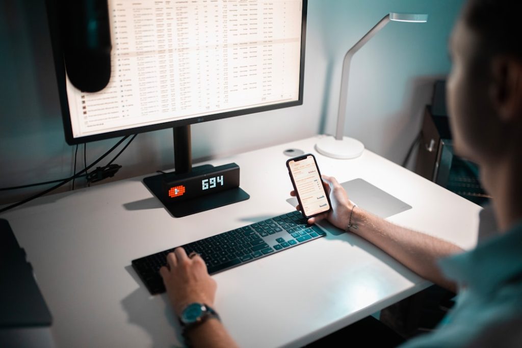 A person works at a dimly lit desk, one hand on the keyboard in front of a monitor full of spreadsheet rows, the other holding a smartphone showing a list of messages.