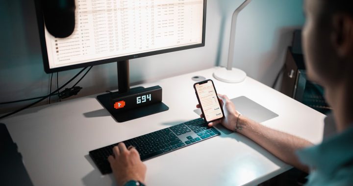 A person works at a dimly lit desk, one hand on the keyboard in front of a monitor full of spreadsheet rows, the other holding a smartphone showing a list of messages.