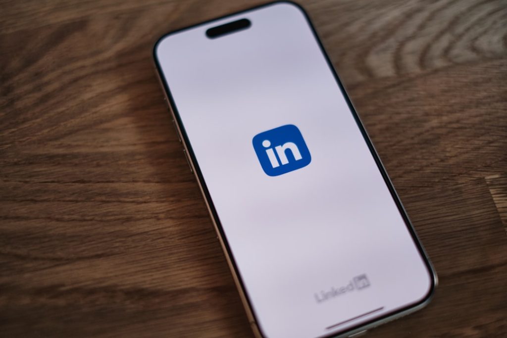 A smartphone lying face-up on a wooden desk, its screen showing the blue LinkedIn app logo on a pale background with the LinkedIn wordmark near the bottom edge.