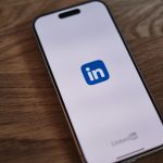 A smartphone lying face-up on a wooden desk, its screen showing the blue LinkedIn app logo on a pale background with the LinkedIn wordmark near the bottom edge.