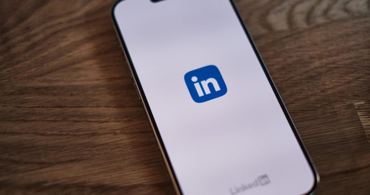 A smartphone lying face-up on a wooden desk, its screen showing the blue LinkedIn app logo on a pale background with the LinkedIn wordmark near the bottom edge.
