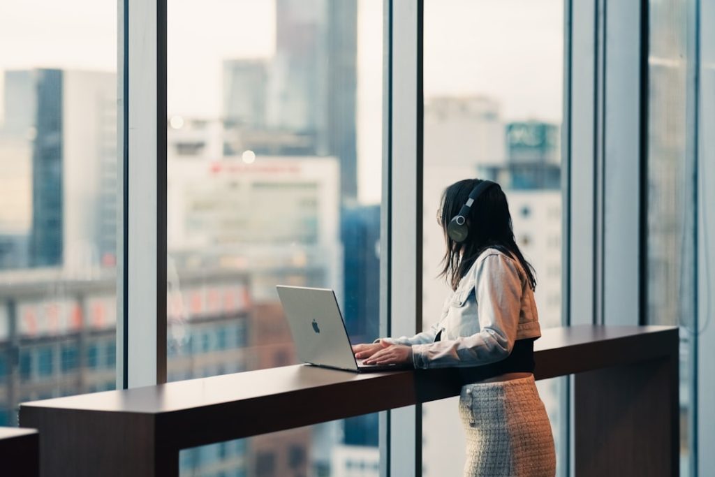 A person in a denim jacket and over-ear headphones stands at a high counter by a floor-to-ceiling window, working on a laptop, with a blurred city skyline behind them.