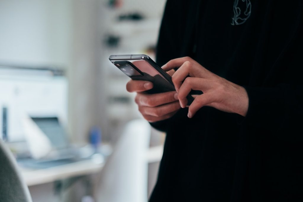 Close-up of a person in a dark sweatshirt holding a smartphone in both hands and typing with their thumbs, standing beside a white desk where a laptop and monitor sit blurred in the background.