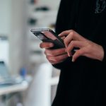 Close-up of a person in a dark sweatshirt holding a smartphone in both hands and typing with their thumbs, standing beside a white desk where a laptop and monitor sit blurred in the background.