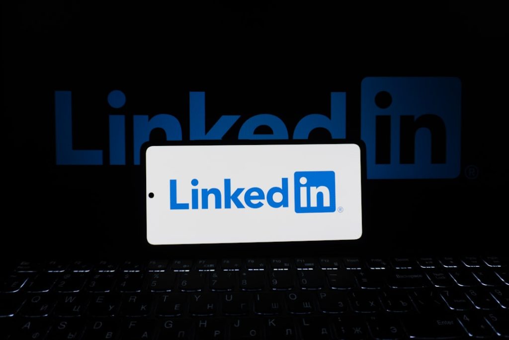 A smartphone propped on a backlit laptop keyboard in a dark room, its screen showing the blue and white LinkedIn logo, with a larger glowing LinkedIn logo on a display behind it.