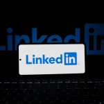 A smartphone propped on a backlit laptop keyboard in a dark room, its screen showing the blue and white LinkedIn logo, with a larger glowing LinkedIn logo on a display behind it.