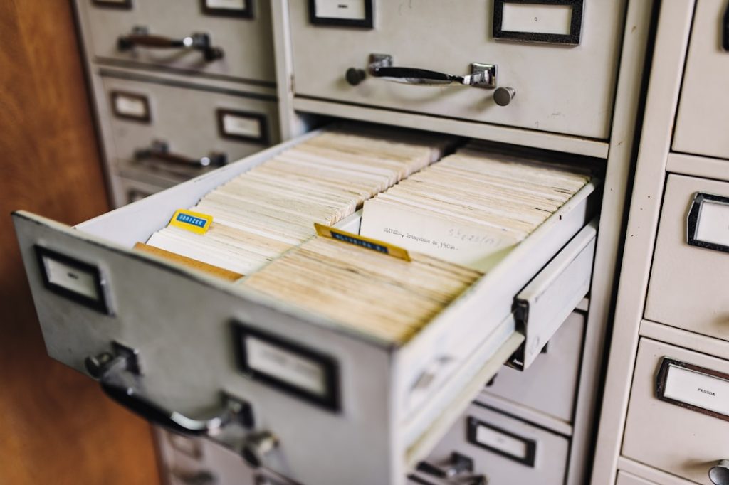An open drawer of a grey metal card catalog packed tight with rows of worn, yellowing index cards, set into a wall of matching filing cabinet drawers.