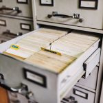 An open drawer of a grey metal card catalog packed tight with rows of worn, yellowing index cards, set into a wall of matching filing cabinet drawers.