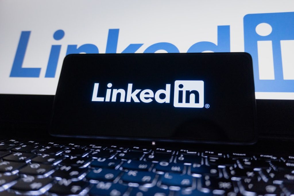 A smartphone resting on a laptop keyboard, its screen showing the white LinkedIn wordmark, with a larger LinkedIn logo glowing on the display behind it.