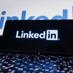 A smartphone resting on a laptop keyboard, its screen showing the white LinkedIn wordmark, with a larger LinkedIn logo glowing on the display behind it.