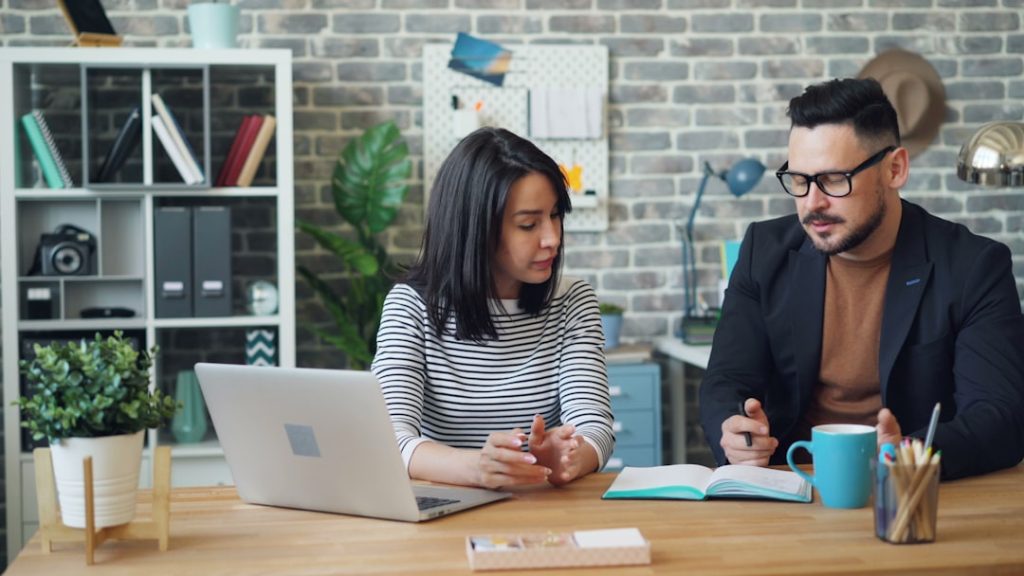 Two coworkers sitting side by side at a wooden desk in a brick-walled office, one working at an open laptop while the other writes in an open notebook next to a teal mug and a cup of pencils.