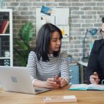 Two coworkers sitting side by side at a wooden desk in a brick-walled office, one working at an open laptop while the other writes in an open notebook next to a teal mug and a cup of pencils.