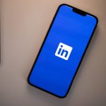 A smartphone lying face up on a pale grey desk next to the corner of a silver laptop, its screen showing the LinkedIn app splash screen with the white in logo centred on a solid blue background.