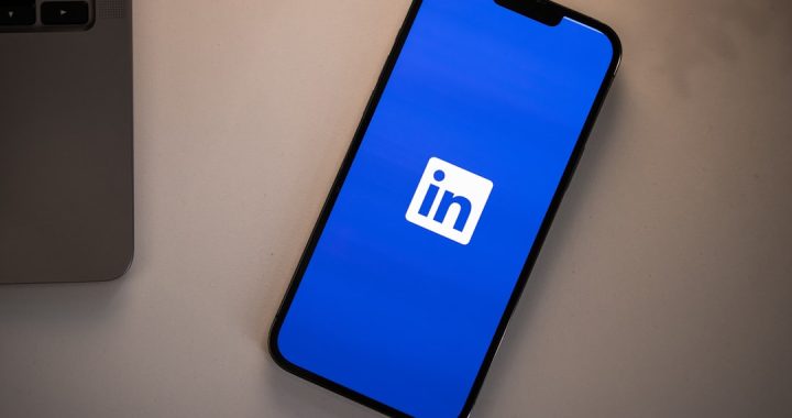 A smartphone lying face up on a pale grey desk next to the corner of a silver laptop, its screen showing the LinkedIn app splash screen with the white in logo centred on a solid blue background.