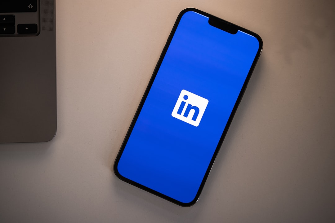 A smartphone lying face up on a pale grey desk next to the corner of a silver laptop, its screen showing the LinkedIn app splash screen with the white in logo centred on a solid blue background.