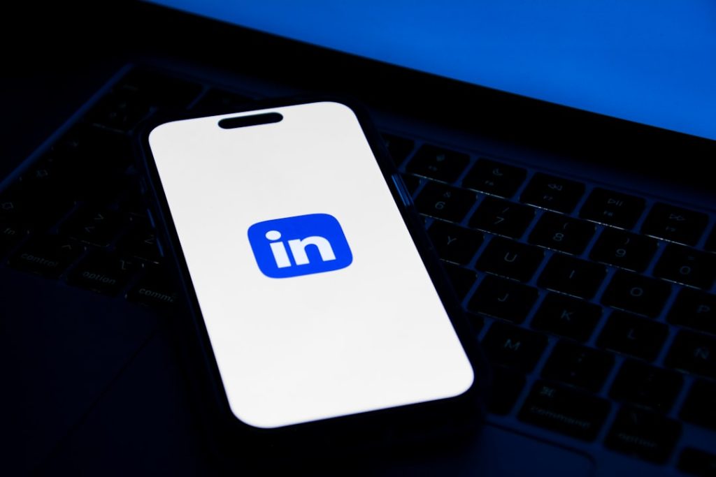 A smartphone resting on a laptop keyboard, its screen showing the blue and white LinkedIn logo, lit against a dark desk.