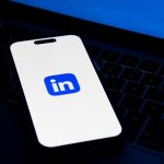 A smartphone resting on a laptop keyboard, its screen showing the blue and white LinkedIn logo, lit against a dark desk.