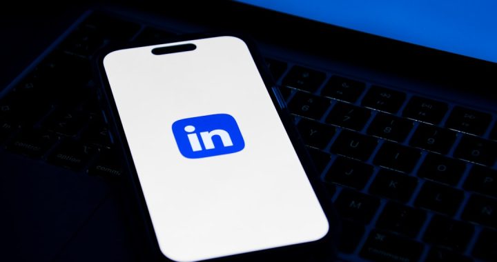 A smartphone resting on a laptop keyboard, its screen showing the blue and white LinkedIn logo, lit against a dark desk.