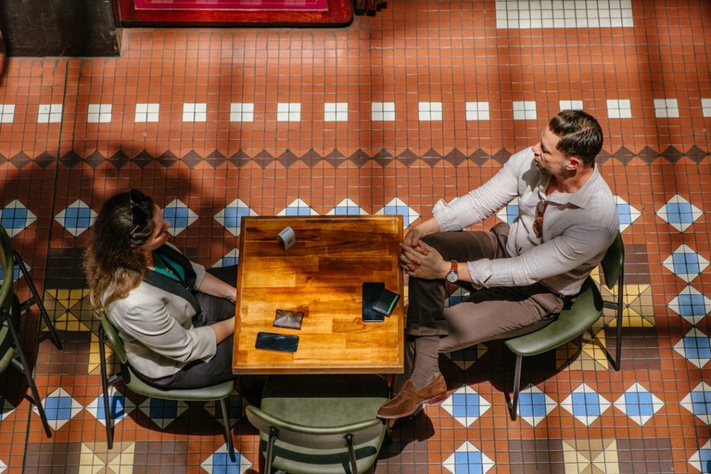 Overhead view of two business-casual people sitting across a small wooden cafe table mid-conversation, phones and wallets on the table, on a patterned tile floor.