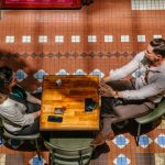 Overhead view of two business-casual people sitting across a small wooden cafe table mid-conversation, phones and wallets on the table, on a patterned tile floor.