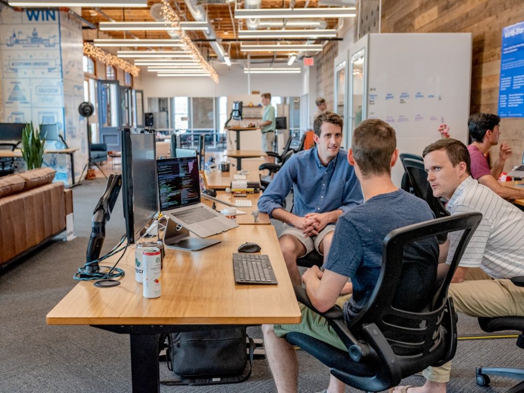 Three colleagues in a small open-plan office sitting together at a long wooden desk in mid-conversation, a monitor, open laptop and keyboard in front of them and a whiteboard covered in notes along the far wall.