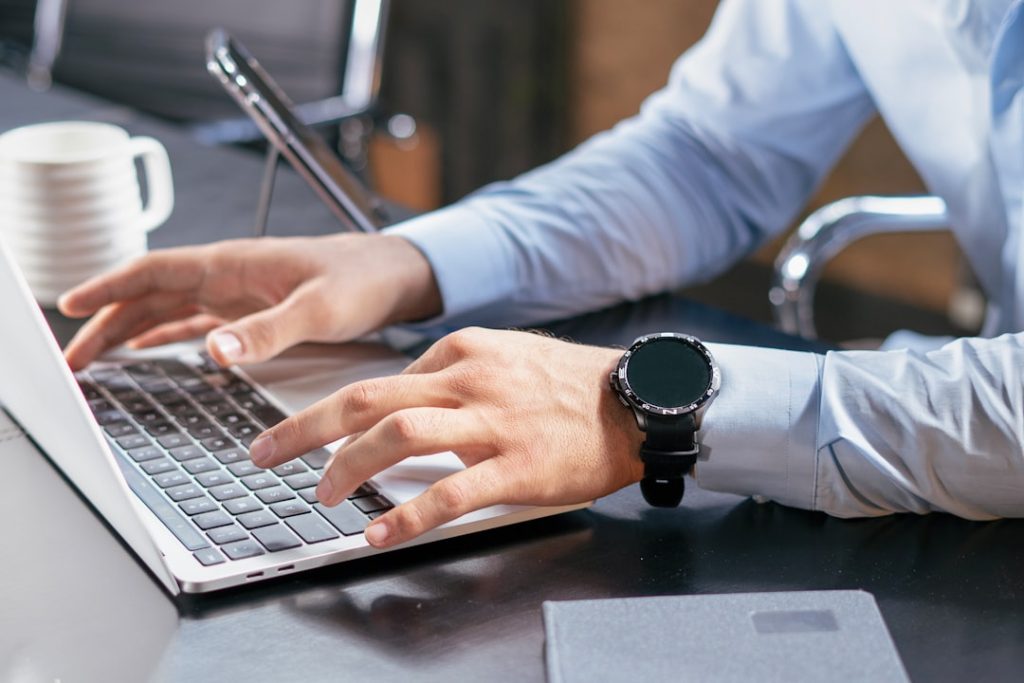 Close view of a person in a light blue dress shirt typing with both hands on a silver laptop at a dark desk, a black smartwatch on their left wrist and a white coffee mug beside the keyboard.