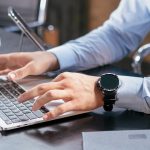Close view of a person in a light blue dress shirt typing with both hands on a silver laptop at a dark desk, a black smartwatch on their left wrist and a white coffee mug beside the keyboard.
