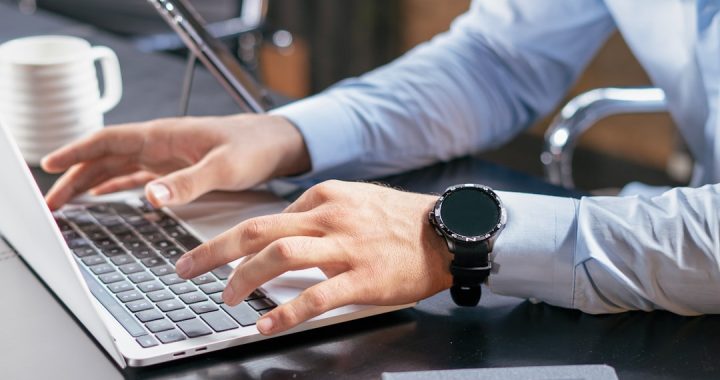 Close view of a person in a light blue dress shirt typing with both hands on a silver laptop at a dark desk, a black smartwatch on their left wrist and a white coffee mug beside the keyboard.
