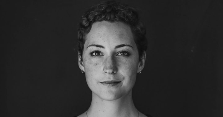 A black and white head-and-shoulders portrait of a person looking straight into the camera against a plain dark background, with a slight closed-mouth smile.