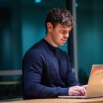 A person in a dark navy sweater sits at a wooden desk in a dimly lit office at night, typing on an open laptop with a glass wall behind them.