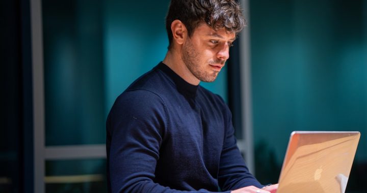 A person in a dark navy sweater sits at a wooden desk in a dimly lit office at night, typing on an open laptop with a glass wall behind them.
