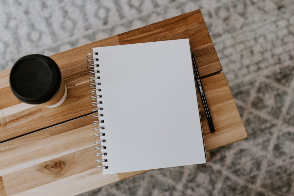 A blank spiral-bound notebook lying open on a light wood table, a pencil resting on the wood beside it and a takeaway coffee cup with a black lid at the top left, photographed from directly above.