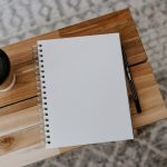 A blank spiral-bound notebook lying open on a light wood table, a pencil resting on the wood beside it and a takeaway coffee cup with a black lid at the top left, photographed from directly above.