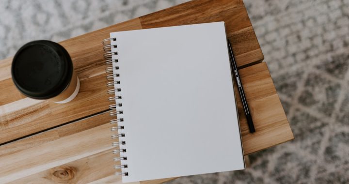 A blank spiral-bound notebook lying open on a light wood table, a pencil resting on the wood beside it and a takeaway coffee cup with a black lid at the top left, photographed from directly above.
