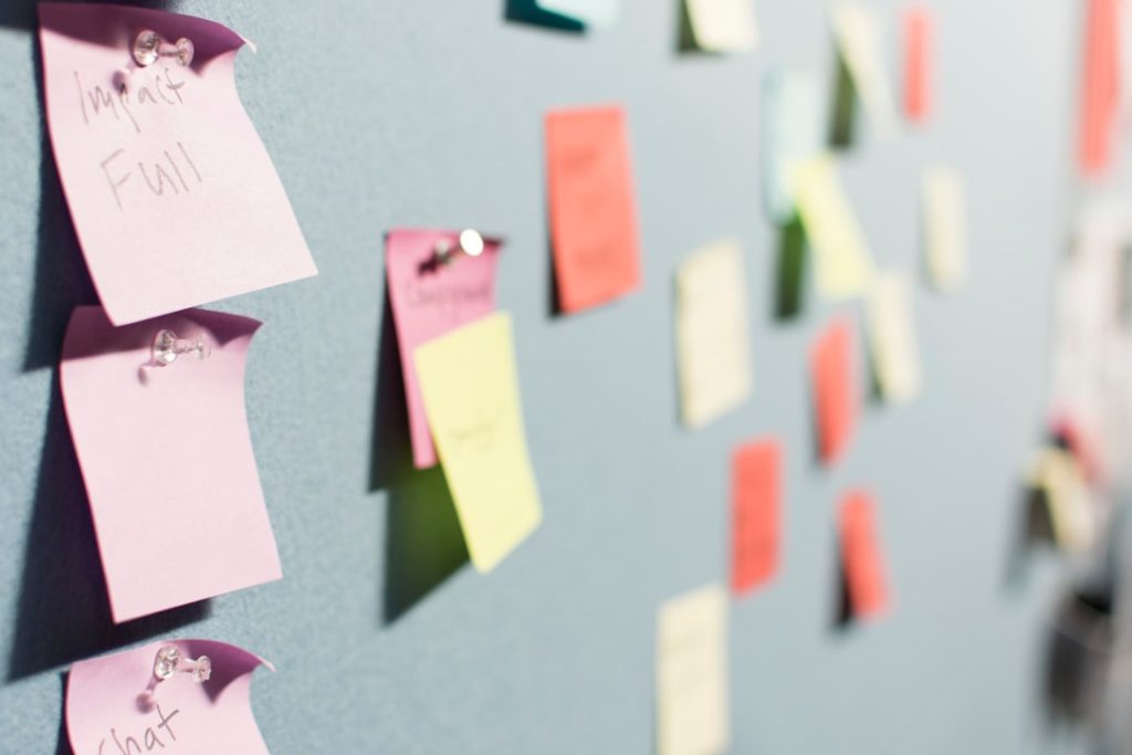 A pale grey board covered in pinned paper sticky notes in pink, coral, yellow and mint, photographed from a sharp angle so the three handwritten pink notes in the left foreground are in focus and the rest of the notes recede into blur.