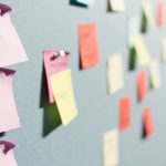 A pale grey board covered in pinned paper sticky notes in pink, coral, yellow and mint, photographed from a sharp angle so the three handwritten pink notes in the left foreground are in focus and the rest of the notes recede into blur.