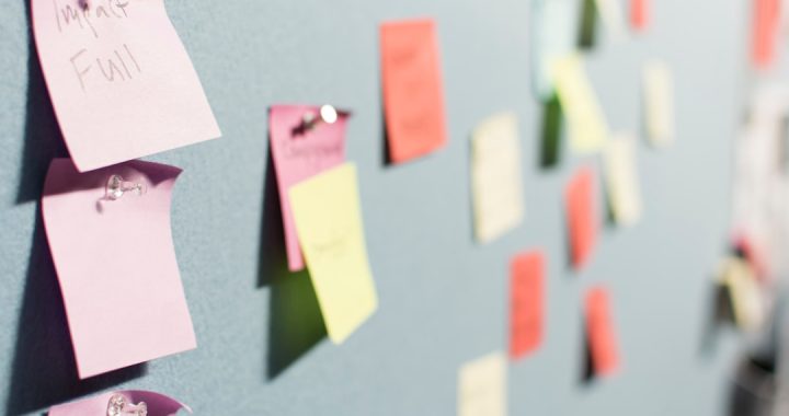 A pale grey board covered in pinned paper sticky notes in pink, coral, yellow and mint, photographed from a sharp angle so the three handwritten pink notes in the left foreground are in focus and the rest of the notes recede into blur.