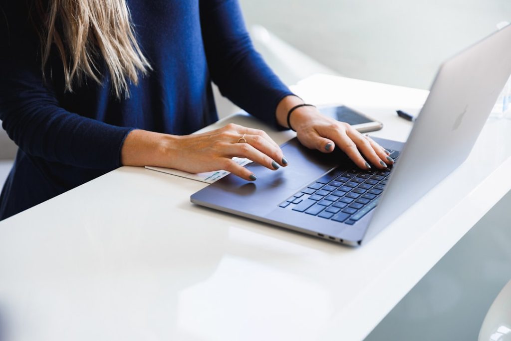 A person in a navy sweater typing on a laptop at a white desk, with a phone and a notepad beside them.
