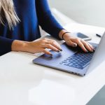 A person in a navy sweater typing on a laptop at a white desk, with a phone and a notepad beside them.