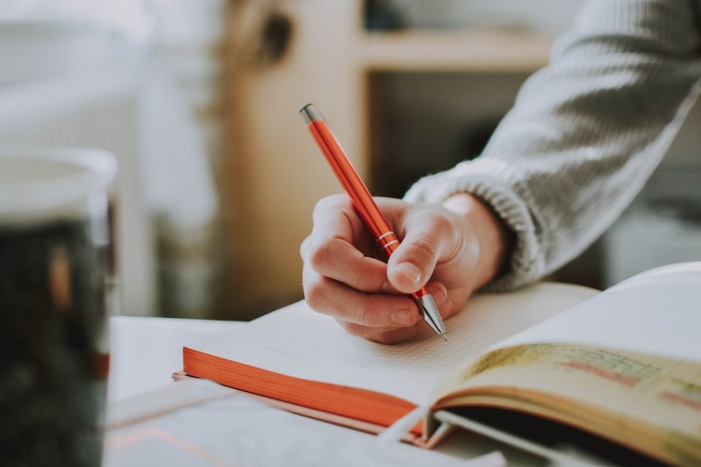 A close-up of a person in a grey knit sweater writing with an orange ballpoint pen in an open grid-ruled notebook, a second open book and a glass beside it on a sunlit desk.