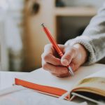 A close-up of a person in a grey knit sweater writing with an orange ballpoint pen in an open grid-ruled notebook, a second open book and a glass beside it on a sunlit desk.