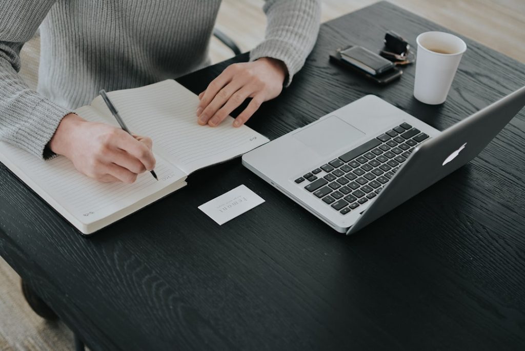 Overhead view of a person in a grey sweater writing notes by hand in a lined notebook at a black desk, next to an open laptop, a paper coffee cup, a wallet and a small business card.