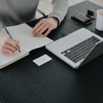 Overhead view of a person in a grey sweater writing notes by hand in a lined notebook at a black desk, next to an open laptop, a paper coffee cup, a wallet and a small business card.