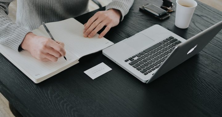 Overhead view of a person in a grey sweater writing notes by hand in a lined notebook at a black desk, next to an open laptop, a paper coffee cup, a wallet and a small business card.
